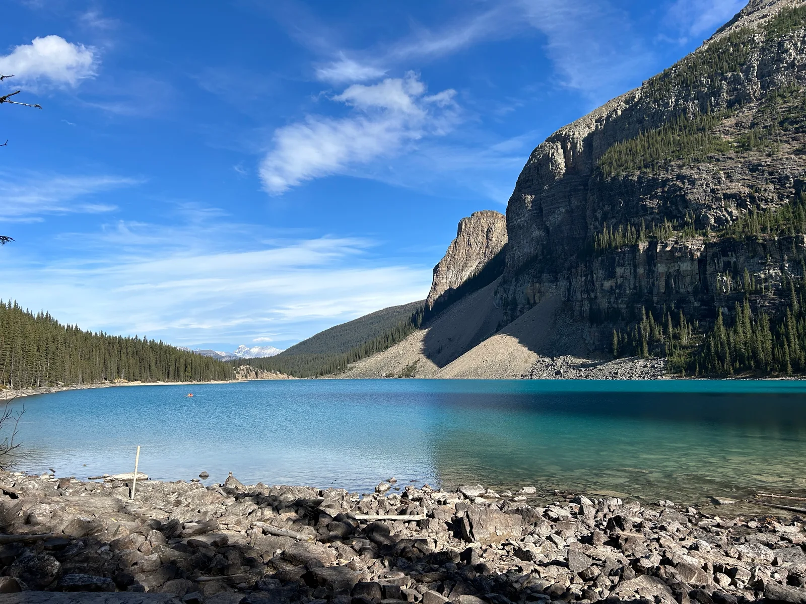 Rocky, driftwood-strewn shoreline at Moraine Lake with turquoise-to-teal water, a lone canoe on the lake, and a jagged peak rising above the far shore under a blue sky