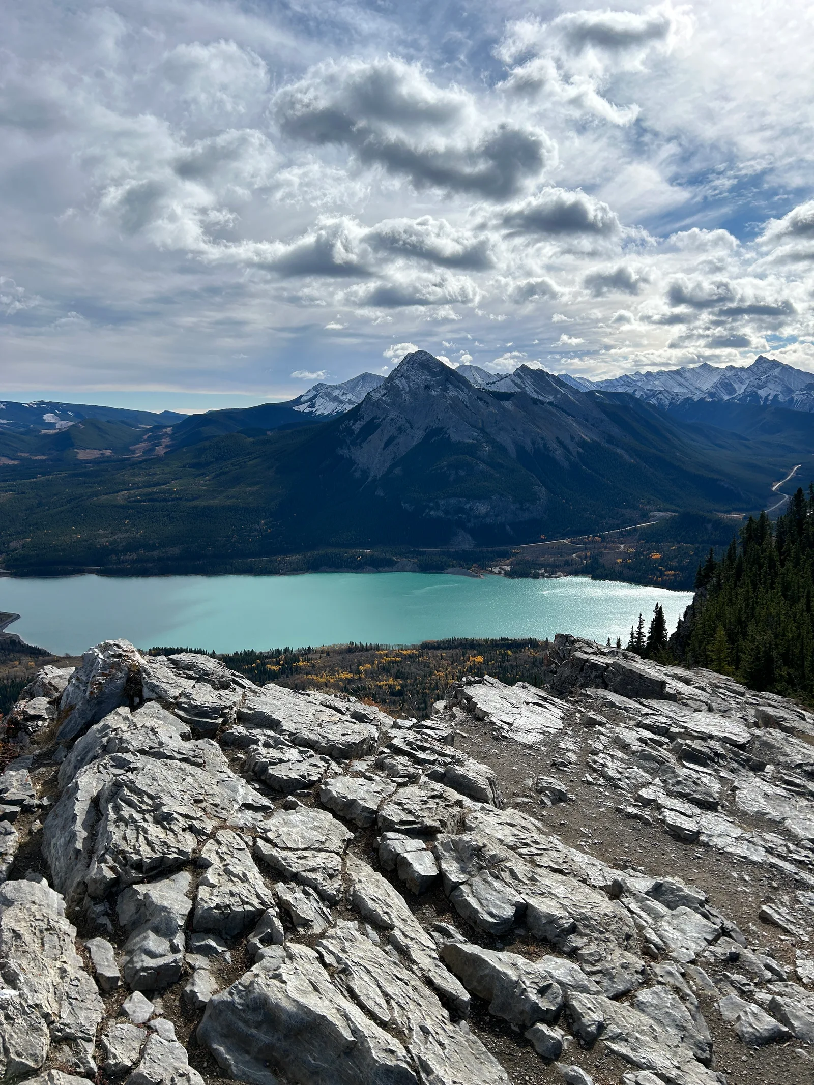Rocky ridge outcrop on Prairie View Trail with a sweeping view down to turquoise Barrier Lake, forest, and mountains under a partly cloudy sky